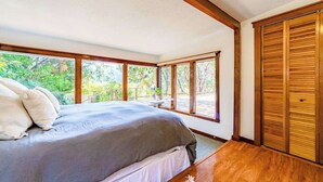 Een slaapkamer met een groot bed, houten vloeren en uitzicht op groen door grote ramen.