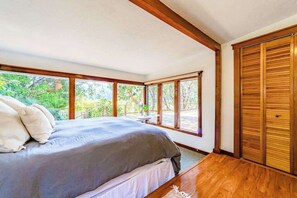 A bedroom with a large bed, wooden flooring, and a view of greenery through large windows. - La Casa Del Rio - Serene Riverside Getaway Surrounded by Nature (Salyer)