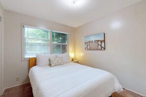 A bedroom with a bed, a window with blinds, a nightstand with a lamp, and a painting on the wall. - Dean Getaway - Peaceful Coastal Escape for the Whole Family (Eureka)