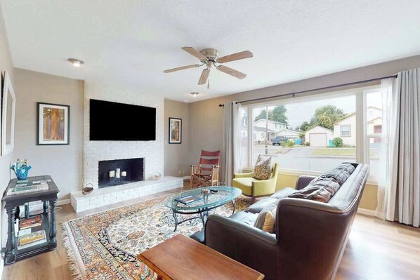 A living room with a fireplace, a ceiling fan, a sofa, and a large window. - Dean Getaway - Peaceful Coastal Escape for the Whole Family (Eureka)