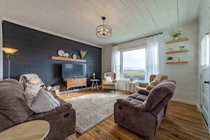 A living room with a dark wall, wooden floor, a television, two sofas, a coffee table, and a view through a window. - 1858 Homestead - Historic Country Retreat with Modern Comforts (Ferndale)