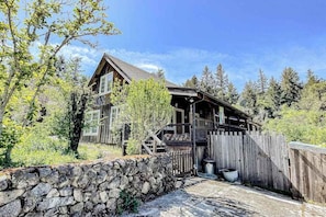 A wooden cabin with a stone wall and a wooden fence, surrounded by trees.
