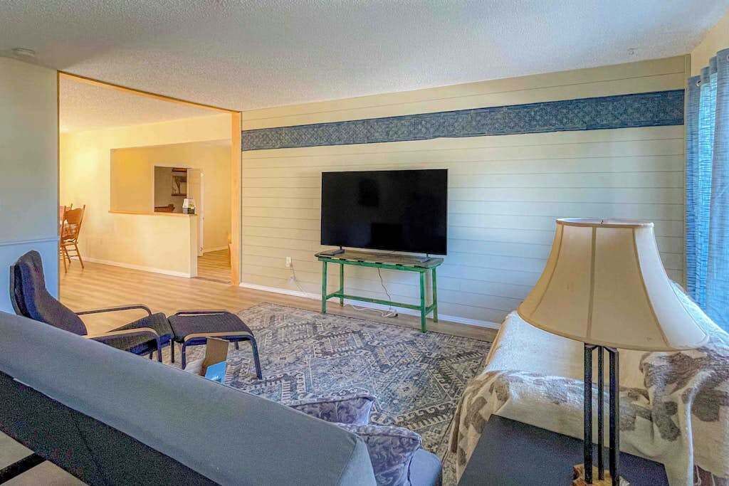Ein Wohnzimmer mit einer Couch, Sesseln, einem Fernseher auf einem Schrank, einer Lampe und einem gemusterten Teppich.
