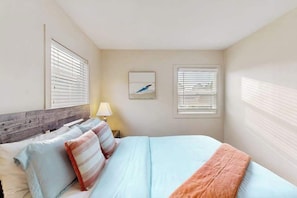 A bedroom with a bed, pillows, a nightstand, a lamp, and two windows with blinds.