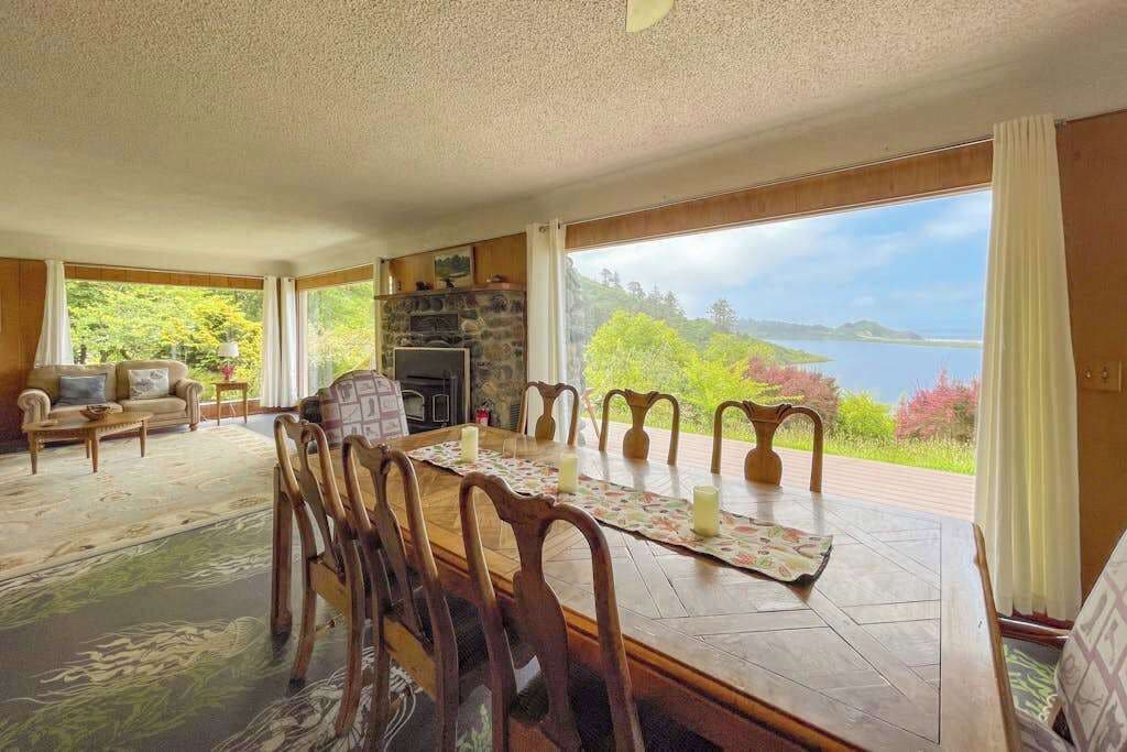 Un comedor con mesa y sillas de madera, chimenea y vistas panorámicas a un lago y vegetación.