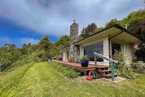 Ein modernes Haus mit einem Stein-Schornstein, einer Terrasse mit Gartenmöbeln und einem üppigen, grünen Hang.