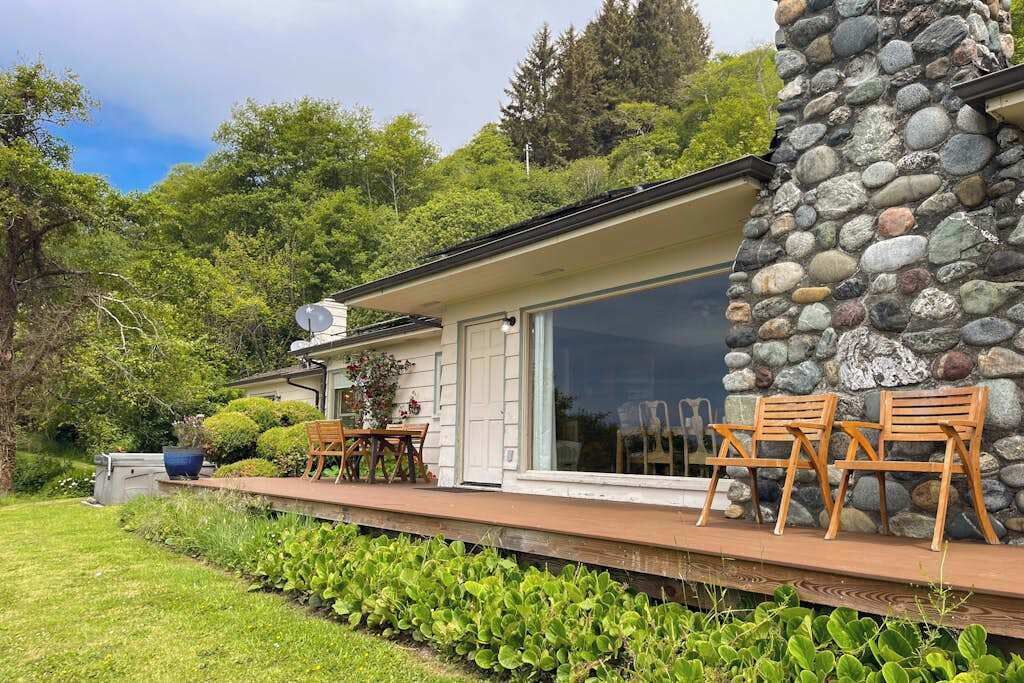 Una casa moderna con fachada de piedra, un porche cubierto con sillas de madera y un ventanal grande.