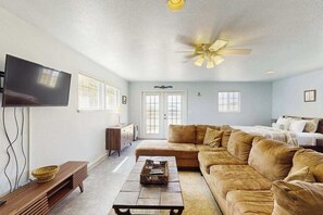 Ein Wohnzimmer mit Ecksofa, Flachbildfernseher, Deckenventilator und einem Fenster mit Jalousien.