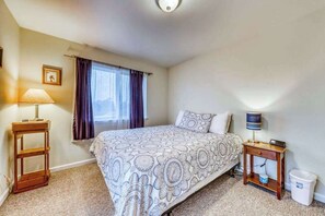 Um quarto com cama, mesa de cabeceira, abajur, quadro na parede e janela com cortinas.