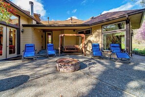 A covered outdoor area with four blue lounge chairs, a fire pit, and a wooden swing structure.