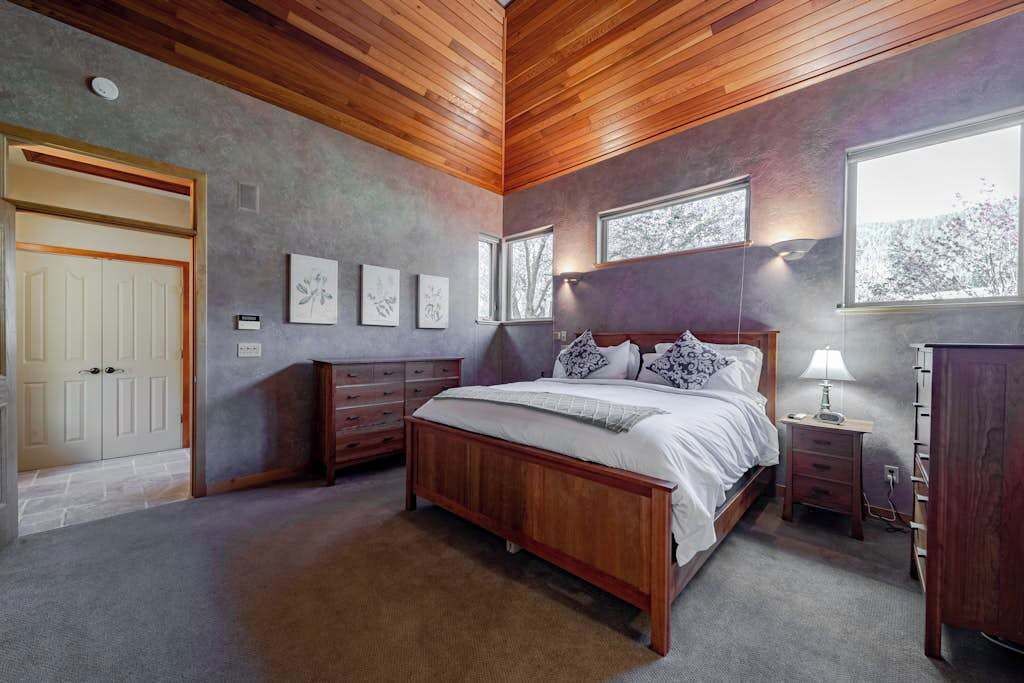 A bedroom with a wooden ceiling, a bed with white bedding, a nightstand with a lamp, and a dresser.