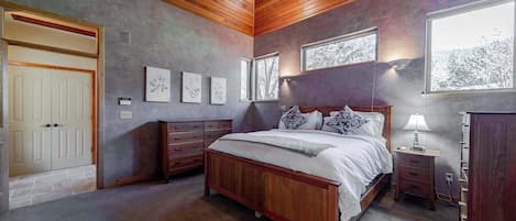 A bedroom with a wooden ceiling, a bed with white bedding, a nightstand with a lamp, and a dresser.