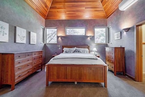 Ein Schlafzimmer mit einem hölzernen Bett, einem Schminktisch und einem Nachttisch. An der Wand hängen gerahmte Bilder und ein Fenster bietet einen Ausblick.
