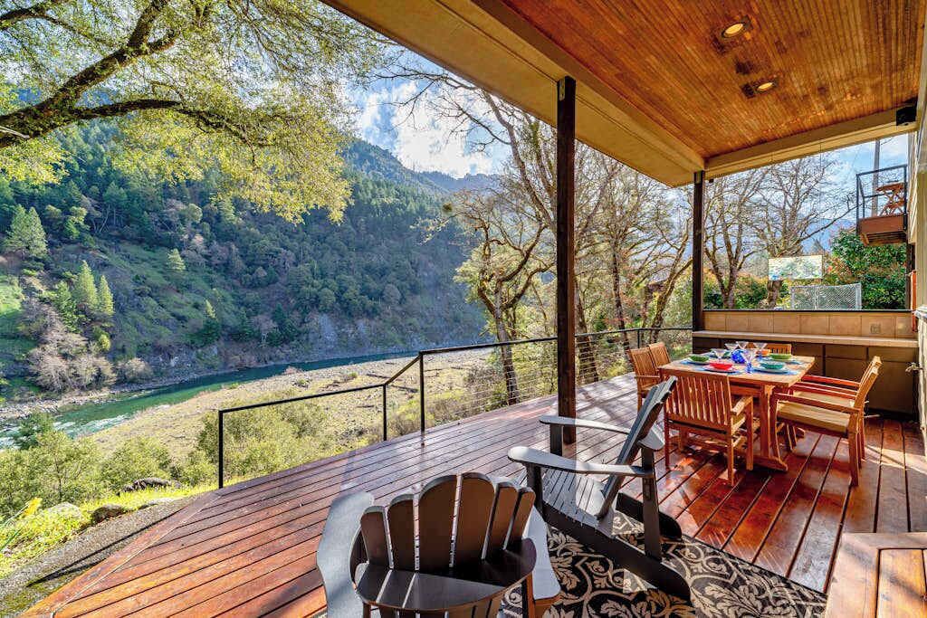 Une terrasse en bois avec une table et des chaises, offrant une vue sur une rivière et des collines boisées.