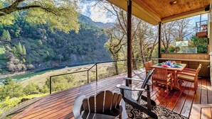 A wooden deck with a table and chairs, overlooking a river and forested hills.