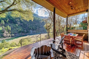 A wooden deck with a table and chairs, overlooking a river and forested hills.