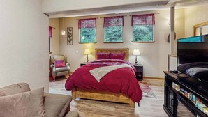A bedroom with a bed, a chair, a TV stand with books, and a window with curtains. - Peaceful Creekside - Tranquil Nature Retreat by the Water (Trinidad)