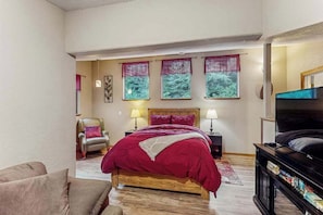 Ein Schlafzimmer mit einem Bett, einem Sessel, einer TV-Kommode mit Büchern und einem Fenster mit Vorhängen.