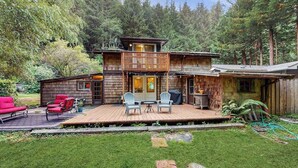 A two-story wooden cabin with a deck, outdoor seating, and a forest backdrop. - Peaceful Creekside - Tranquil Nature Retreat by the Water (Trinidad)