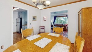 A dining area with a wooden table, chairs, and a chandelier. There is a kitchen visible through an open doorway, and a living room with a sofa and a window. - Eureka Charmer - Cozy Historic Getaway in the Heart of Town (Eureka)