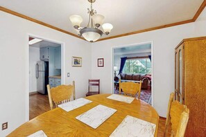 Ein Essbereich mit Holztisch, Stühlen und einer Deckenleuchte. Durch einen offenen Durchgang ist eine Küche zu sehen, und ein Wohnzimmer mit einer Couch und einem Fenster.
