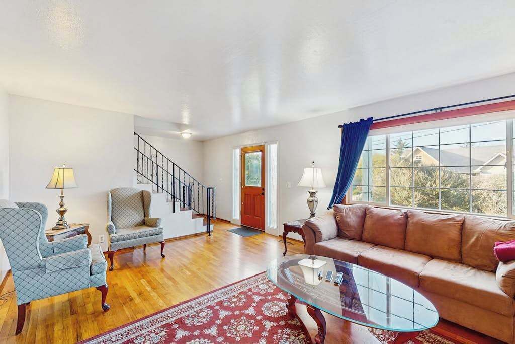 A spacious living room with a wooden floor, a glass coffee table, a sofa, and two armchairs. A staircase is visible in the background.