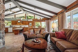 A spacious living area with a leather sofa, a coffee table, and a dining table. There is a kitchen area with a clock and a window overlooking greenery. - Viewcrest Retro Country Home (Orick)