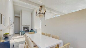 A modern dining area with a white table and chairs, a chandelier, and a view of the kitchen in the background. - Pacific View Escape - Stunning Ocean Views & Coastal Comfort (Crescent City)