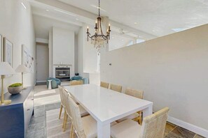 Ein moderner Essbereich mit einem weißen Tisch und Stühlen, einer Kristallleuchter und Blick zur Küche im Hintergrund.