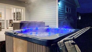 Un jacuzzi con vapor saliendo, rodeado por una terraza de madera y una casa con porche cubierto.