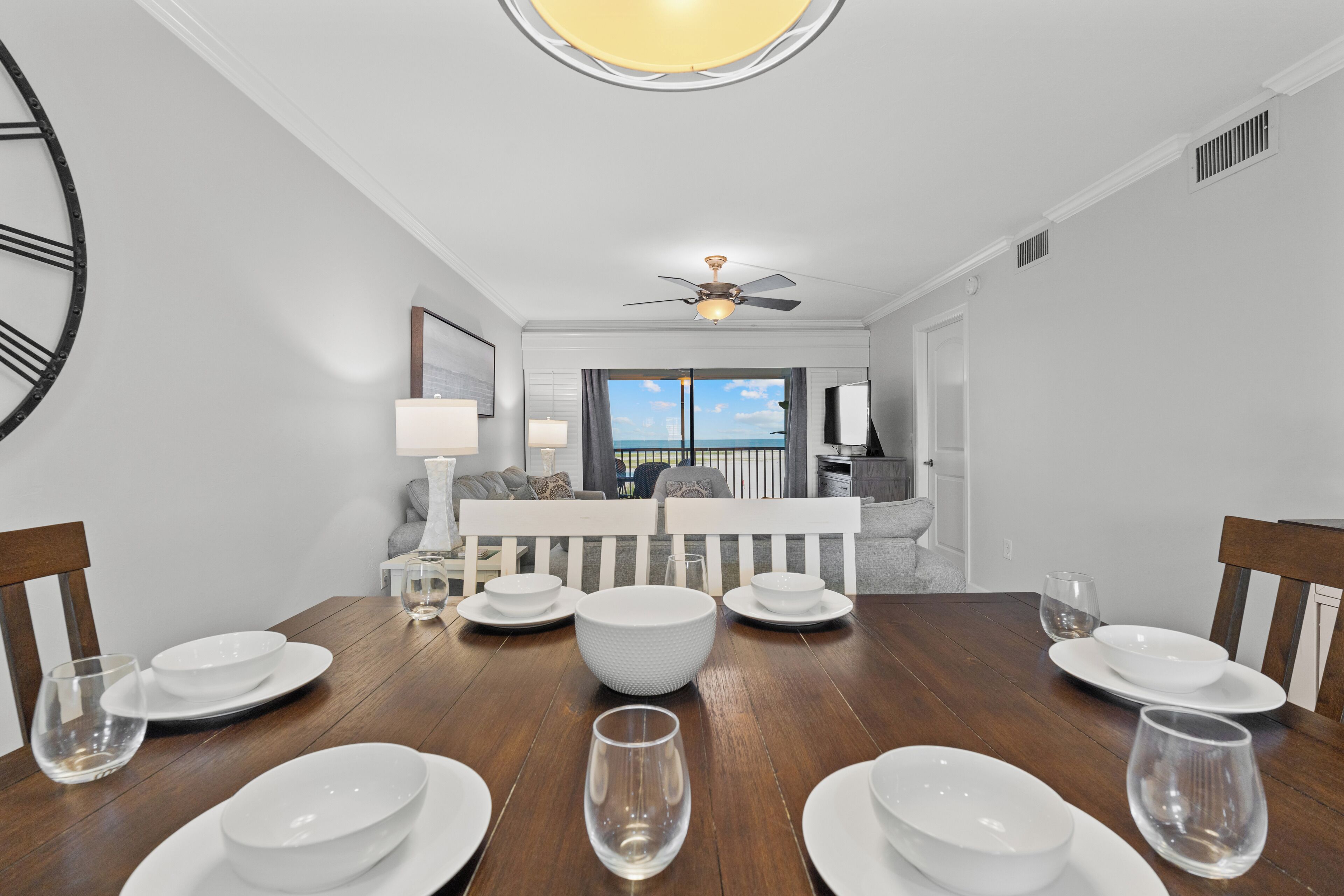 Un coin repas avec une table en bois dressée pour six personnes, un ventilateur de plafond et une vue sur l’océan par une grande fenêtre.