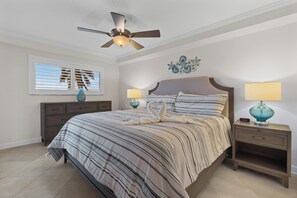 A bedroom with a bed, two bedside tables, a ceiling fan, a window with blinds, and a wall decoration. - ⛱️ Coastal Views | Beachfront Location | Paradise Point–Carlos Pointe #213 🌊 (Fort Myers Beach)