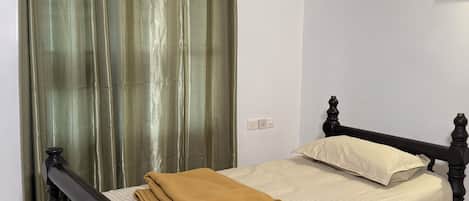Una cama individual con estructura de madera y colchón color beige, una manta marrón doblada encima y una cortina verde entreabierta.