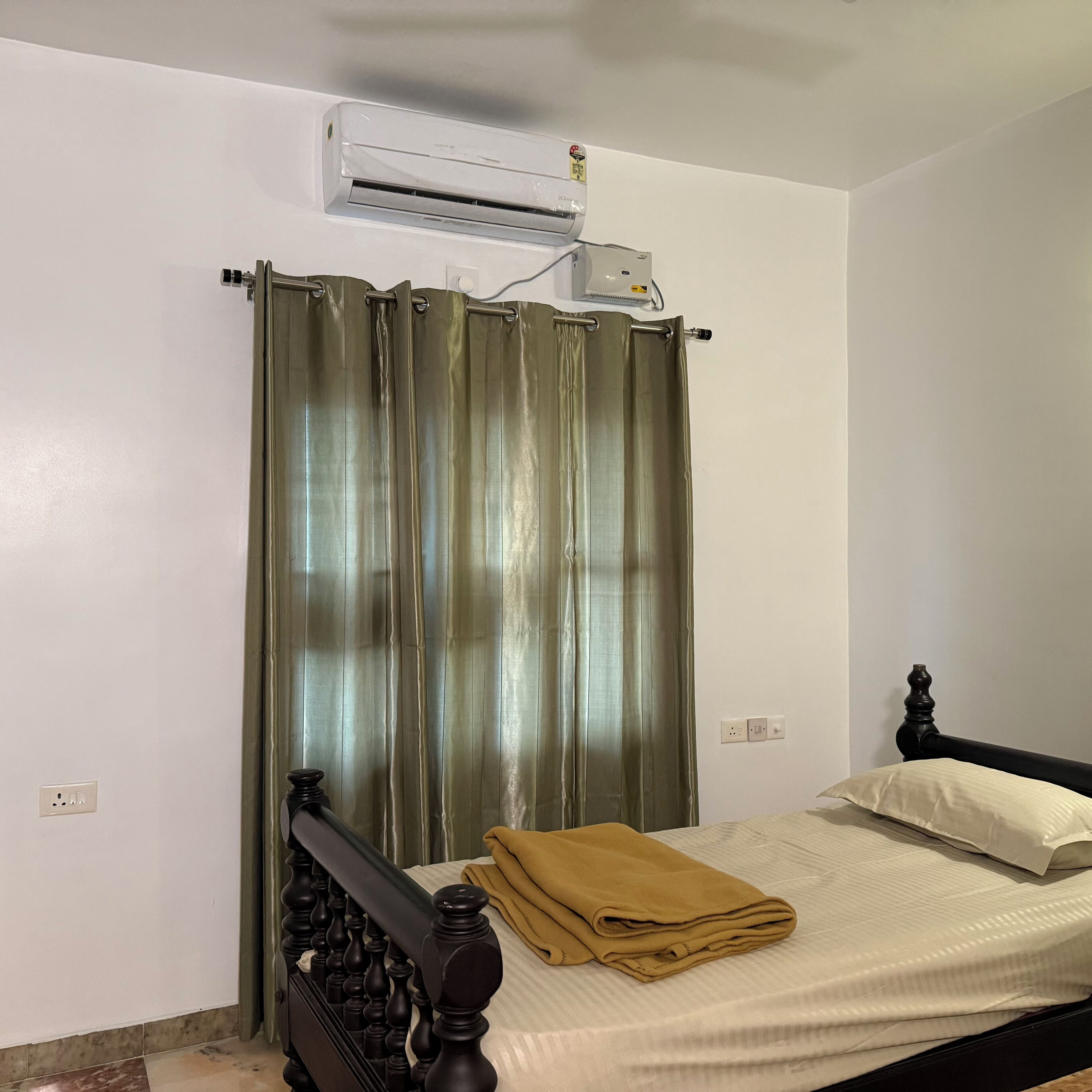 A bedroom with a bed, a folded blanket, a curtain, an air conditioning unit, and a wall socket.