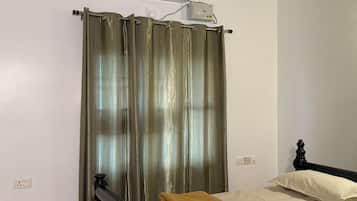 A bedroom with a bed, a folded blanket, a curtain, an air conditioning unit, and a wall socket.