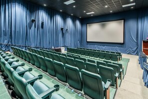 Ein Theater mit Reihen grüner Sitze, einer großen Leinwand und blauen Vorhängen.