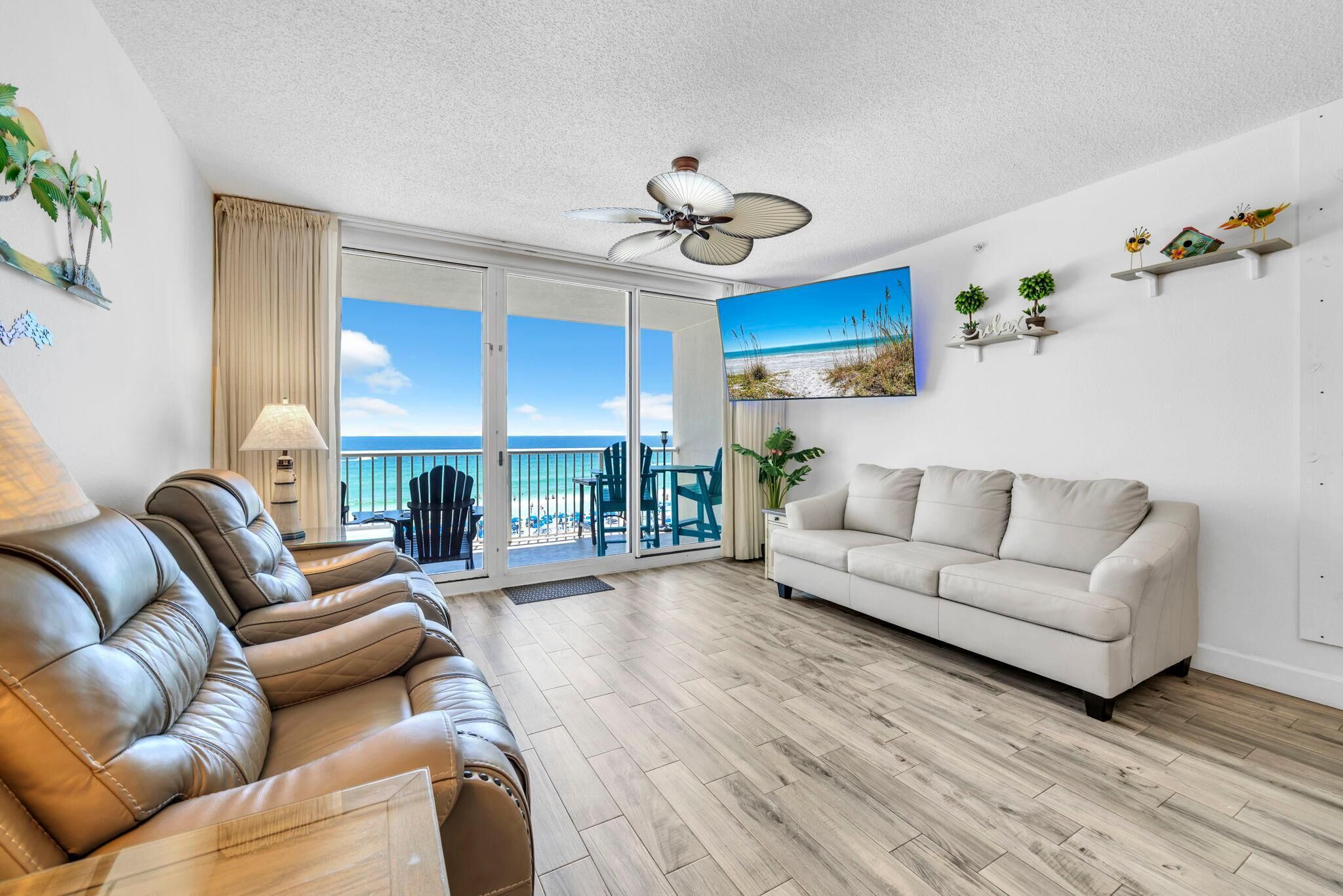 A living room with a leather sofa, a white couch, a ceiling fan, and a view of the beach through the glass door.