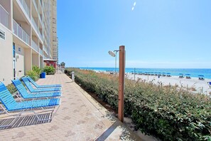 Ein Promenadweg am Strand mit blauen Liegestühlen, einem Holzpfosten mit einer Lampe und Blick auf den Strand und das Meer.