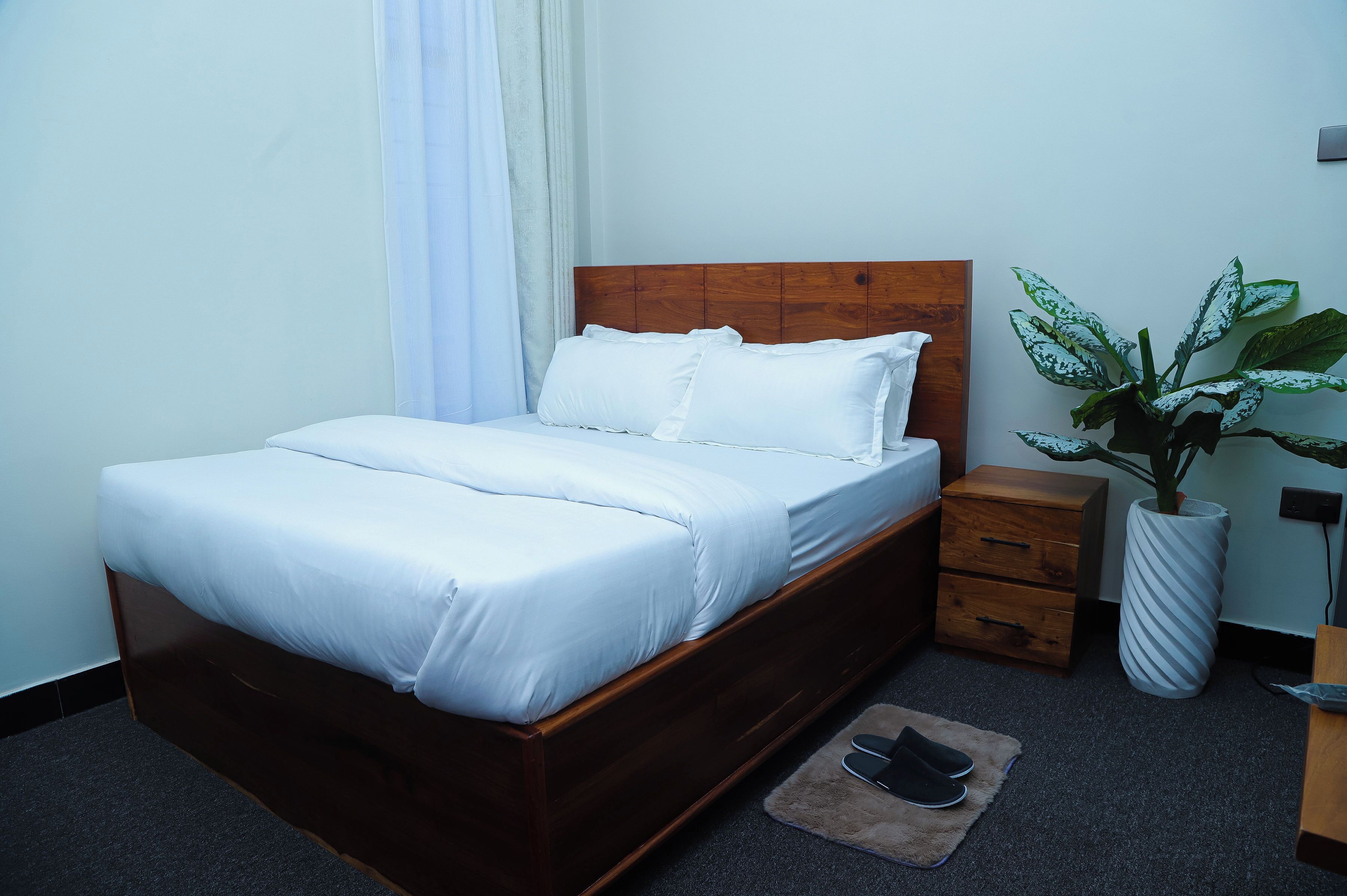 A neatly made bed with white linens, a wooden headboard, a bedside table with drawers, a large potted plant, and a pair of slippers on a rug.