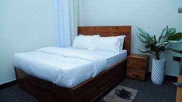 Una cama bien hecha con sábanas blancas, un cabecero de madera, una mesita de noche con cajones, una planta grande en maceta y un par de pantuflas sobre una alfombra.