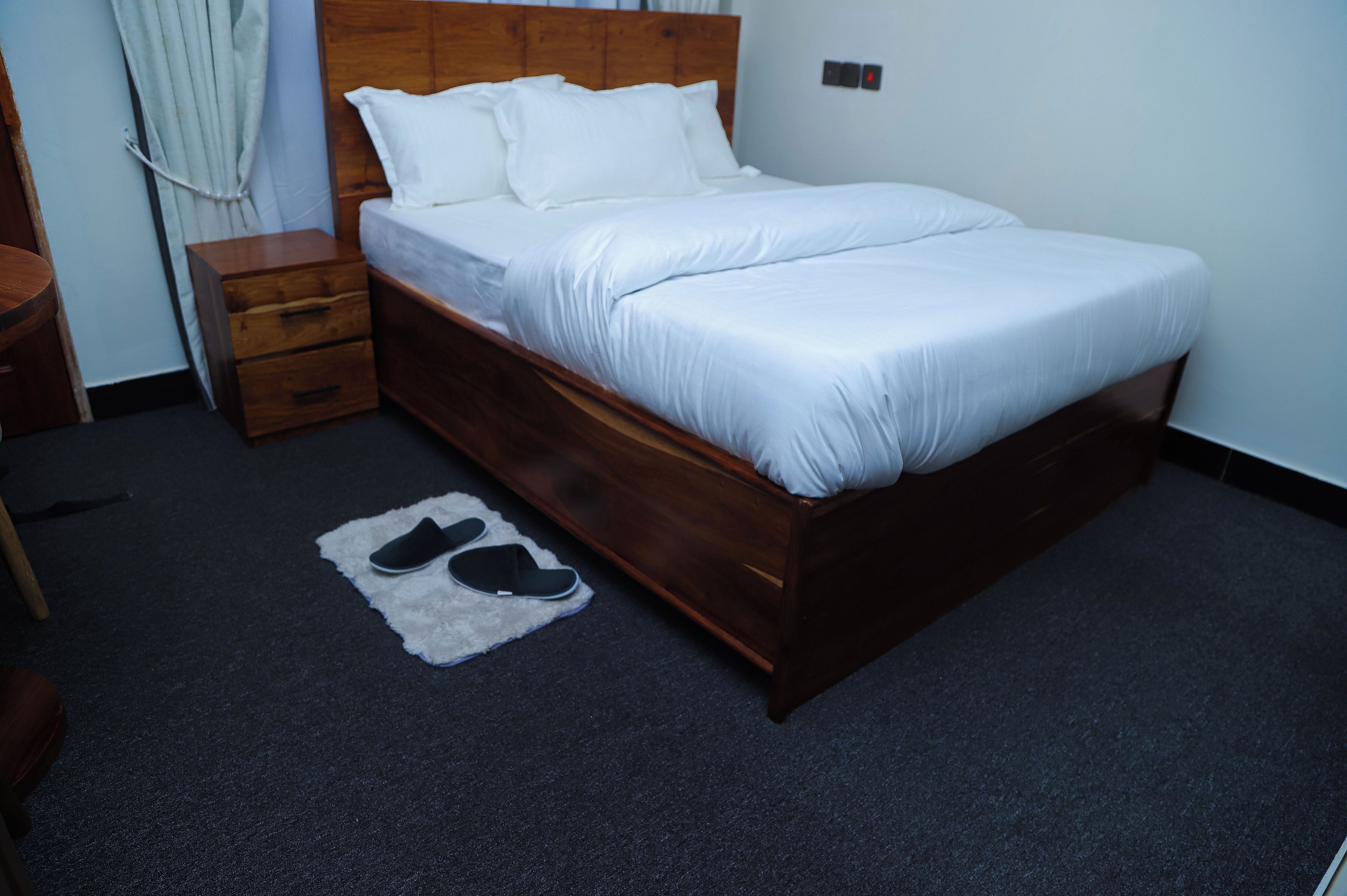 A neatly made bed with white linens in a room with a dark carpet, a wooden nightstand with drawers, and a pair of black slippers on a white rug.