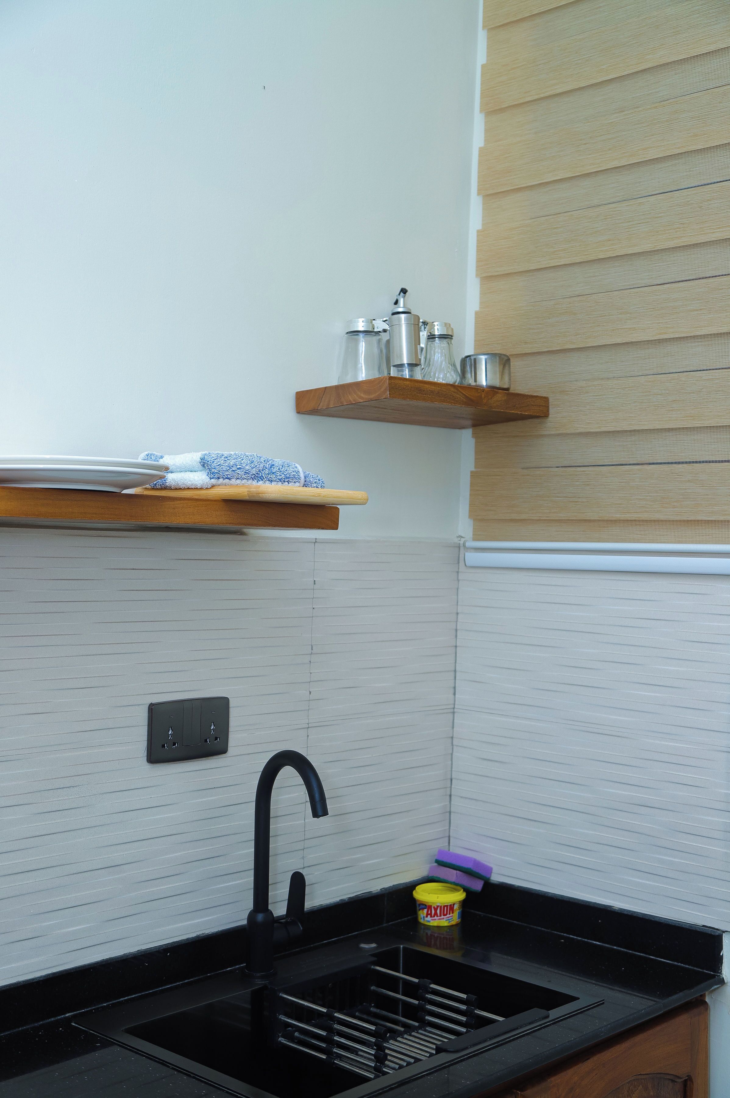 A kitchen with a black sink, a faucet, a wooden shelf with kitchen items, and a tiled backsplash.