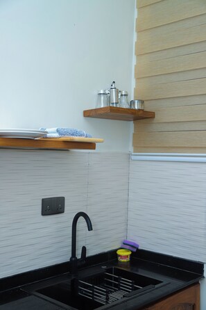 A kitchen with a black sink, a faucet, a wooden shelf with kitchen items, and a tiled backsplash. - Ken Homestay (Arusha)
