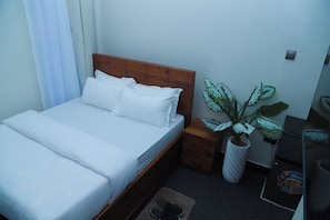 A single bed with white bedding, a wooden headboard, a nightstand with a drawer, a potted plant, and a small table with a mirror and a decorative item. - Ken Homestay (Arusha)