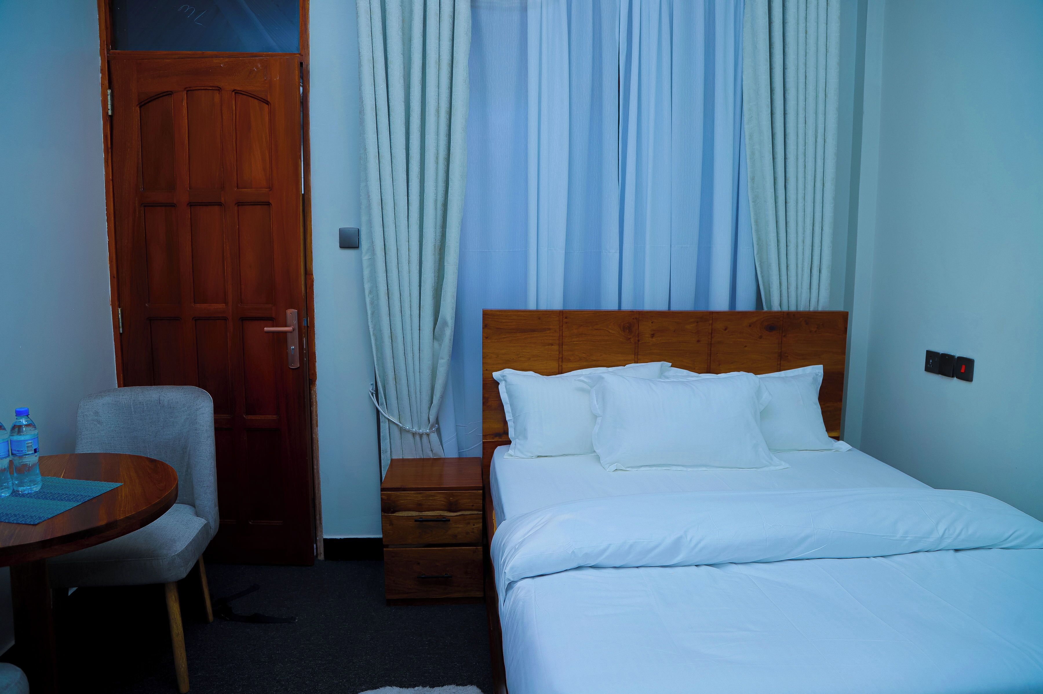 A hotel room with a wooden door, a bed with white bedding, a small wooden nightstand, a chair, and a table with water bottles.