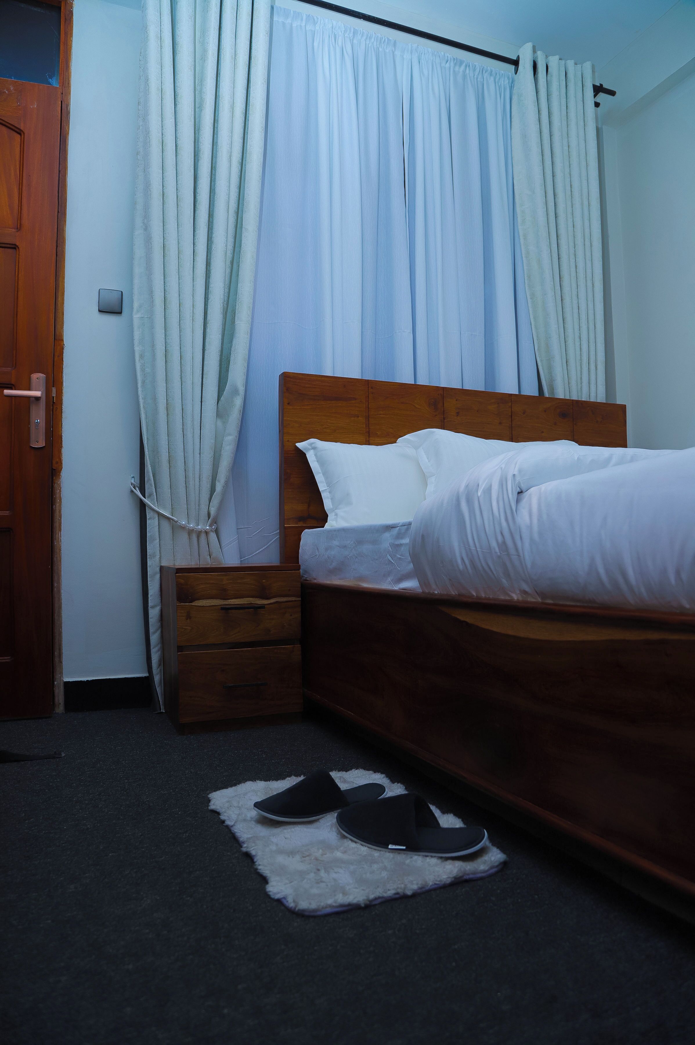 A bedroom with a wooden bed, white bedding, a nightstand, a drawer, and a pair of slippers on a rug.