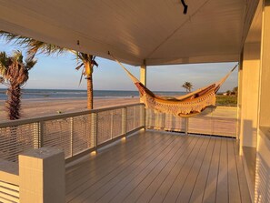 Una terraza de madera con una hamaca, palmeras y vista a la playa.