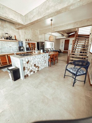 A spacious kitchen with a central island, wooden chairs, a dining table, and a staircase leading to an upper level. - Uba House in Icaraizinho/CE - Beachfront - infinity pool - right on the sand (Icarai)