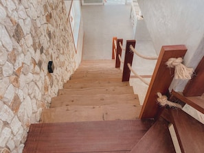 Una escalera de madera con un muro de piedra a un lado y un muro blanco al otro, que sube a un piso superior.
