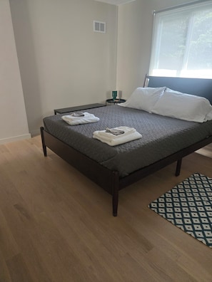 Ein ordentlich bezogenes Bett mit gefalteten Handtüchern, ein Nachttisch mit einem grünen Gegenstand und ein gemustertes Teppich auf Holzboden.